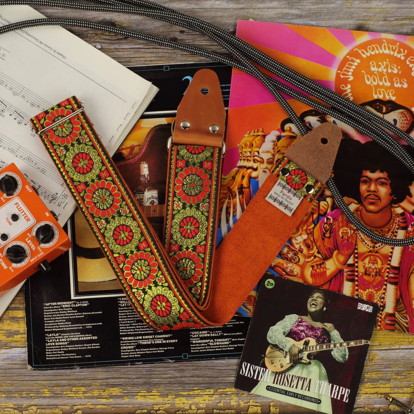 Amber Parade woven guitar strap showing its decorative front pattern and orange suede backing, displayed with vinyl records and guitar accessories.