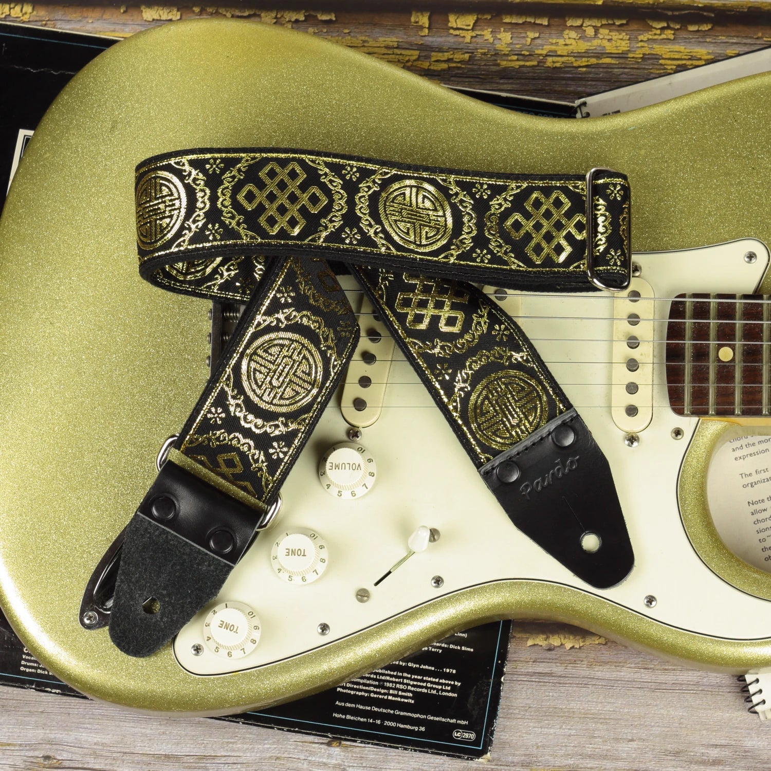 The Medal black and gold guitar strap paired with a Fender Stratocaster Gold Sparkle, showcasing its retro jacquard weave and shimmering gold geometric motifs.