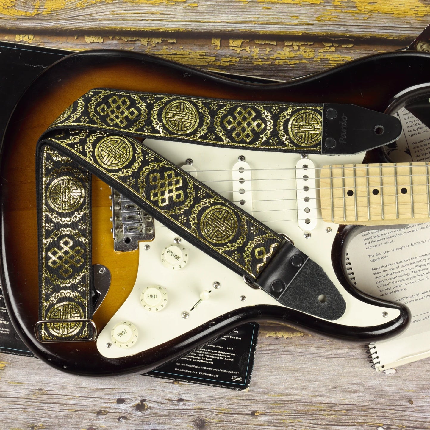 Black and gold Medal guitar strap displayed on a Fender Stratocaster Sunburst, showcasing its shimmering retro jacquard pattern and black leather ends.