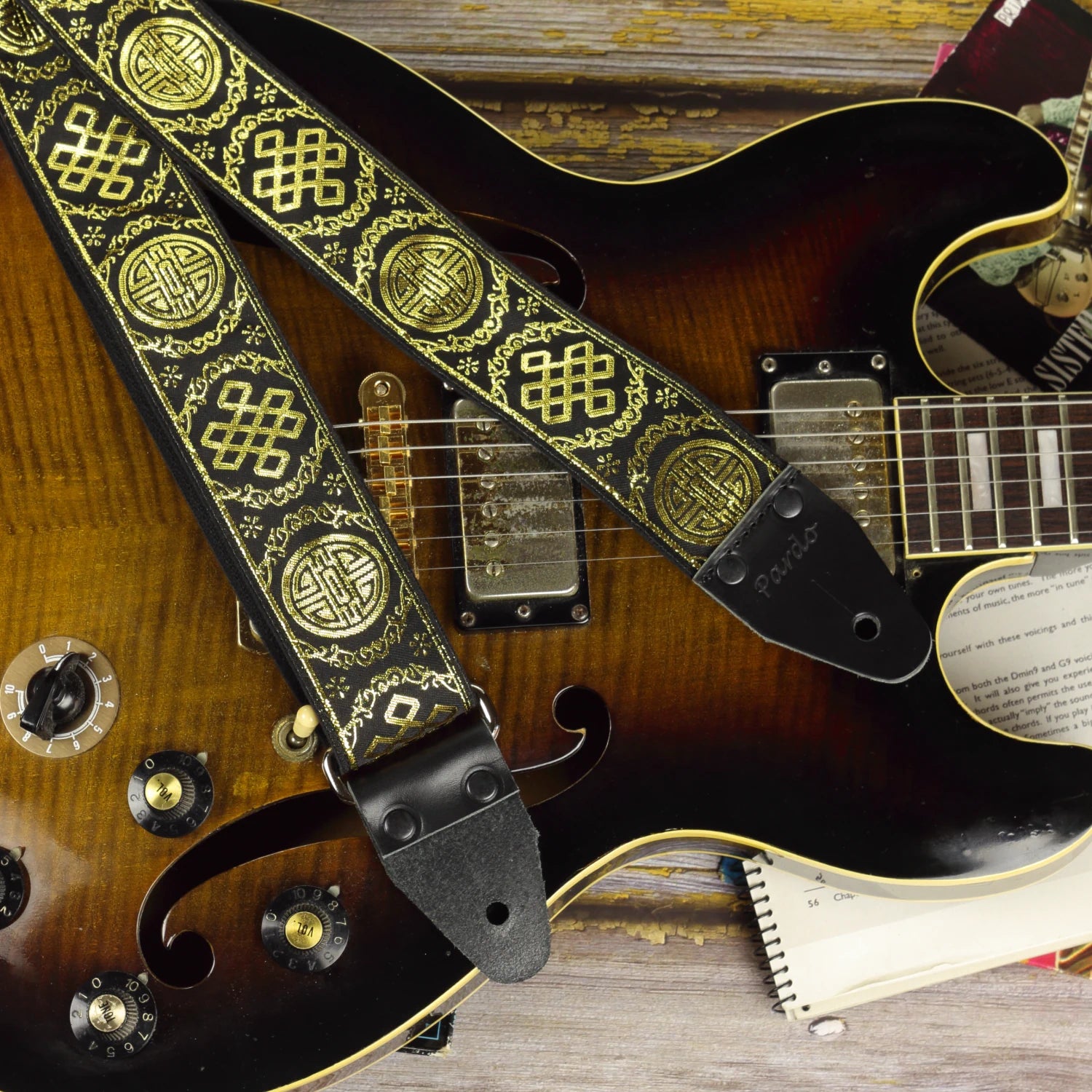 Close-up of the Medal black and gold guitar strap resting on a brown semi-hollow guitar, highlighting its shimmering jacquard weave and genuine leather ends.