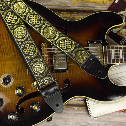 Close-up of the Medal black and gold guitar strap resting on a brown semi-hollow guitar, highlighting its shimmering jacquard weave and genuine leather ends.