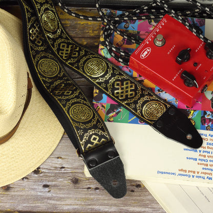 Black and gold Medal guitar strap resting on a guitar tablature book next to a tremolo pedal, showcasing its handcrafted jacquard pattern and genuine leather ends.