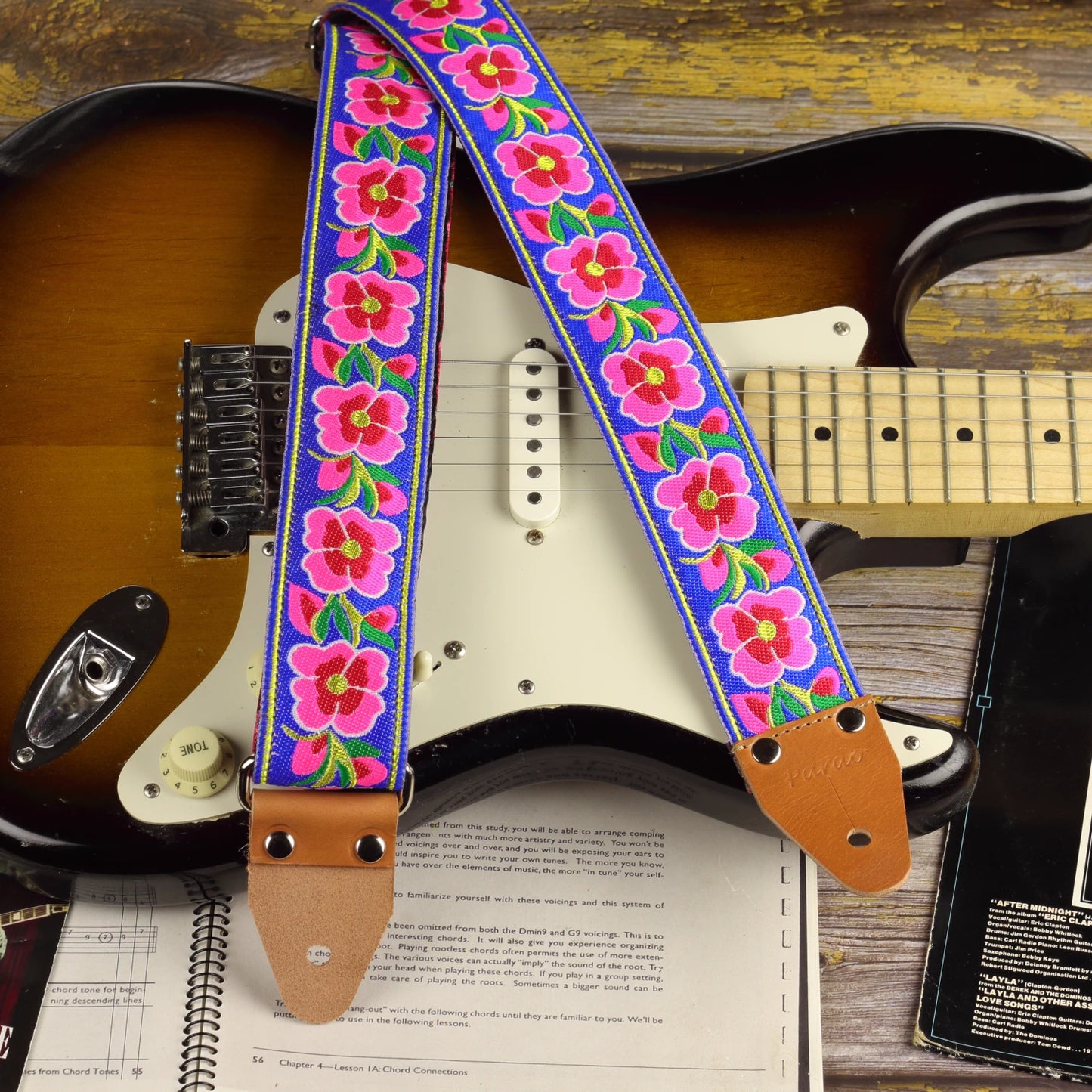 Blue Serenade guitar strap displayed with a sunburst Fender Stratocaster, showing its large pink and red woven motifs on a blue background.