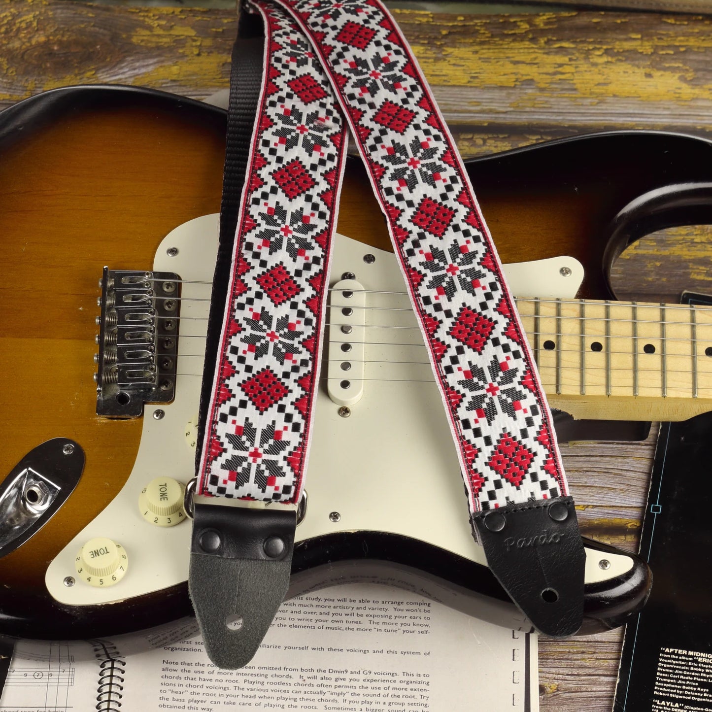 Crimson Compass guitar strap displayed with a sunburst Fender Stratocaster, showing its red, black and ivory geometric pattern.