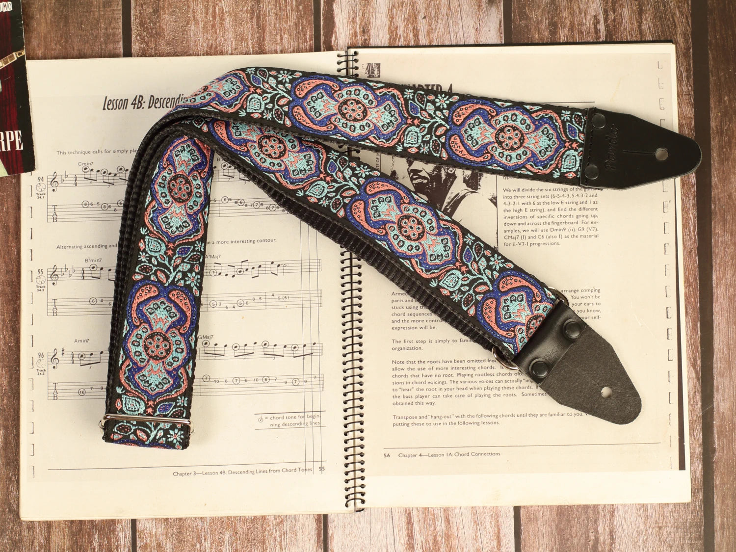 Full-length Blue Mirage handmade guitar strap featuring premium jacquard fabric and genuine leather ends displayed on a vintage guitar sheet music book