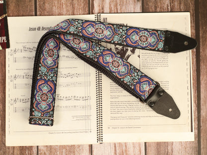 Full-length Blue Mirage handmade guitar strap featuring premium jacquard fabric and genuine leather ends displayed on a vintage guitar sheet music book