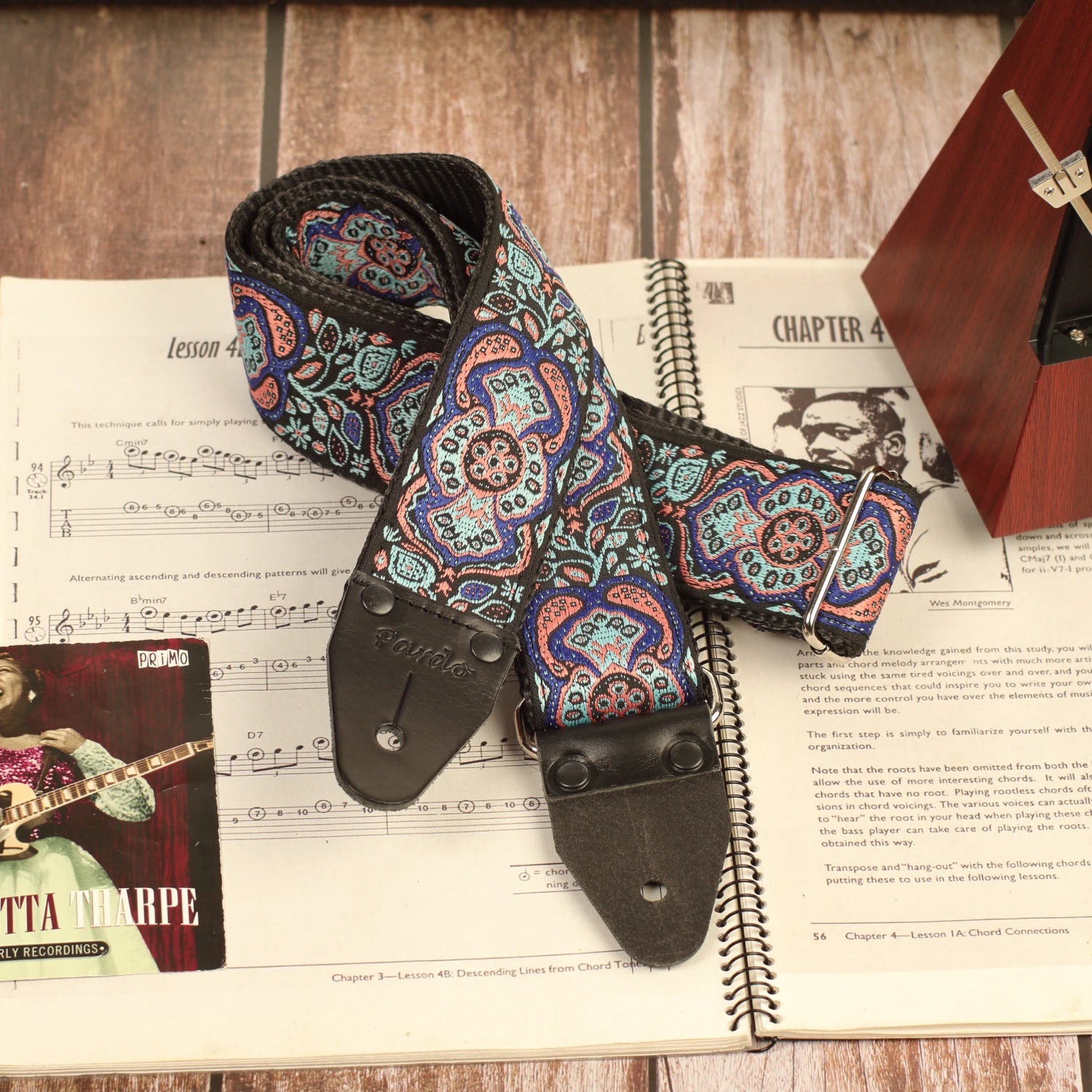 Blue Mirage handmade guitar strap neatly rolled beside a vintage guitar sheet music book and a mechanical metronome