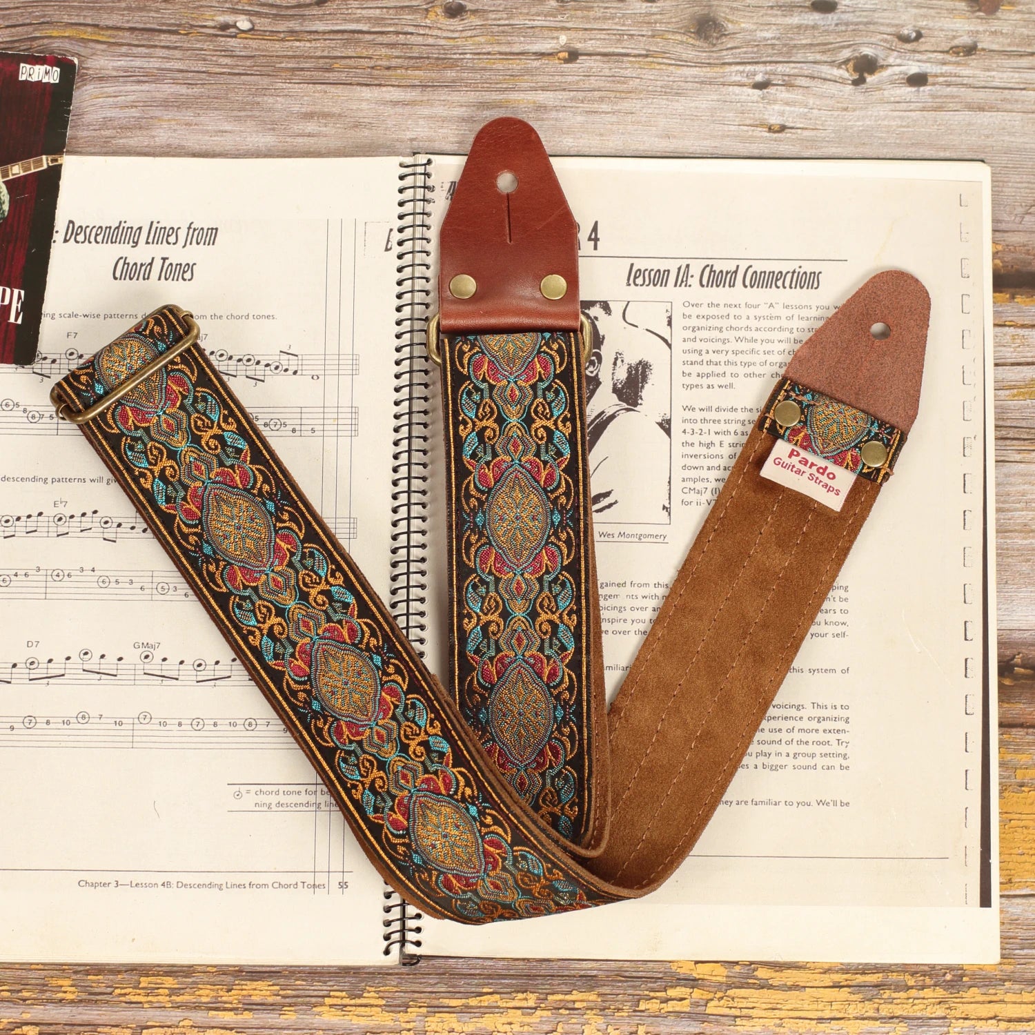 Close-up of the Loyalty multicolored jacquard guitar strap on a sheet music book, highlighting the woven fabric, dark brown suede backing, and handcrafted finish.