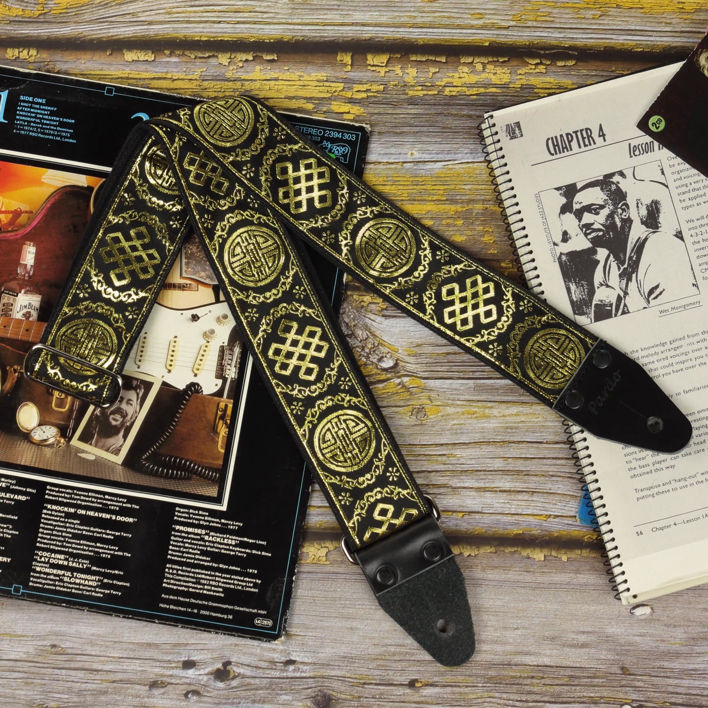 The Medal retro black and gold guitar strap displayed with a sheet music book and an Eric Clapton vinyl record, highlighting its shimmering jacquard weave and handcrafted vintage character.