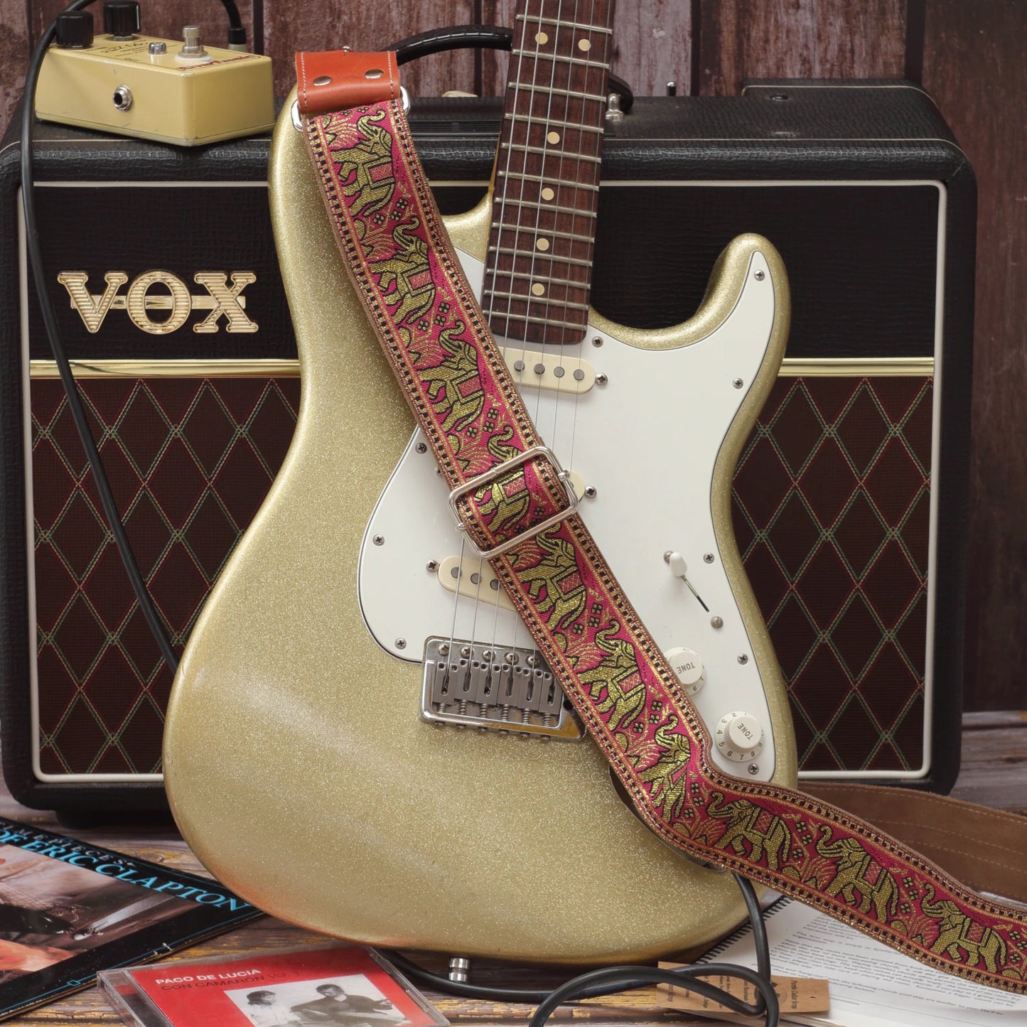 Pink and gold elephant jacquard guitar strap displayed with a Fender Stratocaster and Vox AC10 guitar amplifier