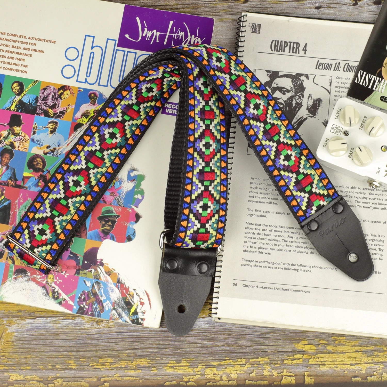 Prism Mosaic guitar strap displayed on a sheet music book, showing its multicolored jacquard pattern and black leather ends.