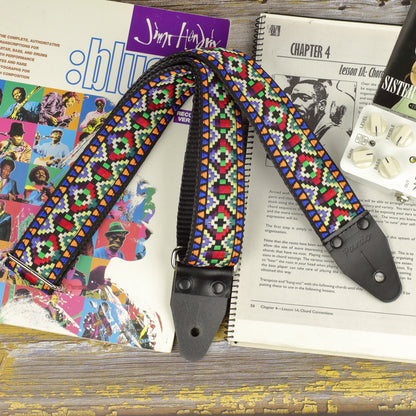 Prism Mosaic guitar strap displayed on a sheet music book, showing its multicolored jacquard pattern and black leather ends.