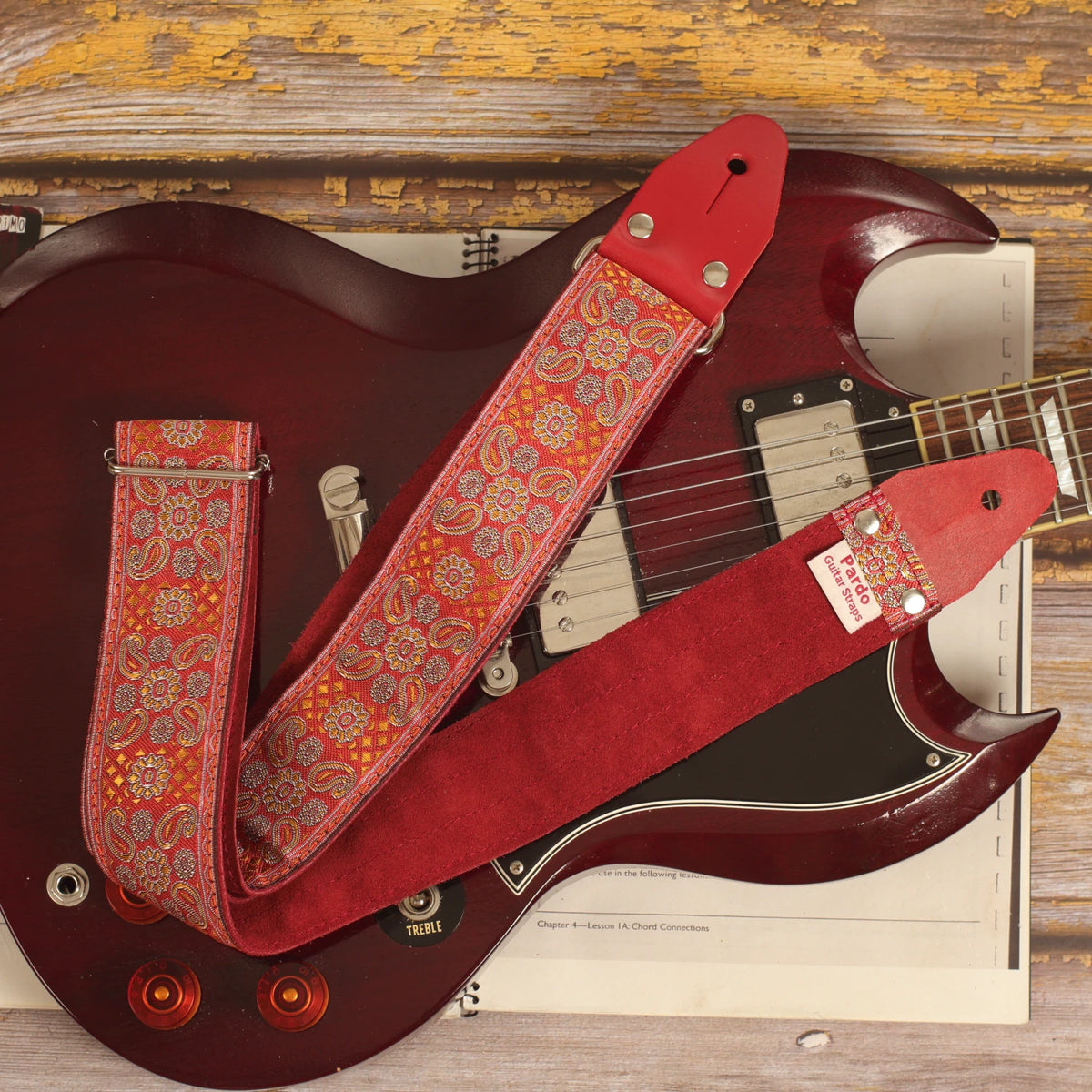 Red Andromeda guitar strap laid over an Edwards SG guitar, showing its red and orange jacquard pattern and burgundy suede backing.