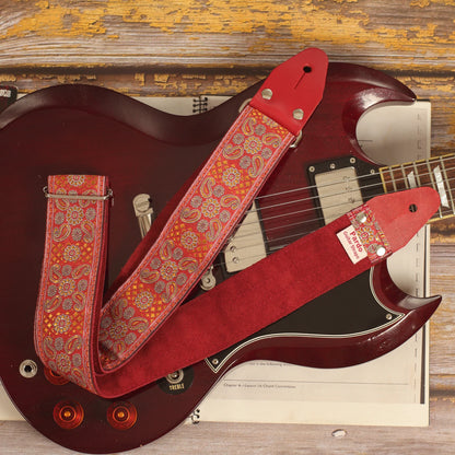 Red Andromeda guitar strap laid over an Edwards SG guitar, showing its red and orange jacquard pattern and burgundy suede backing.