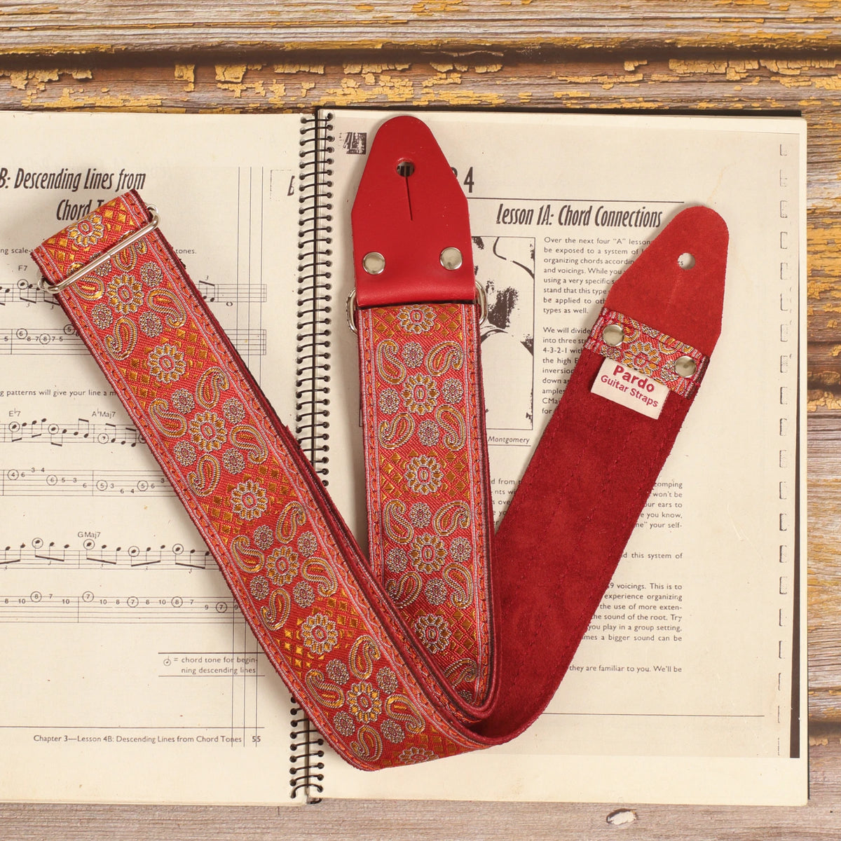 Close-up of the Red Andromeda guitar strap on a sheet music book, showing its red and orange jacquard pattern and burgundy suede backing.