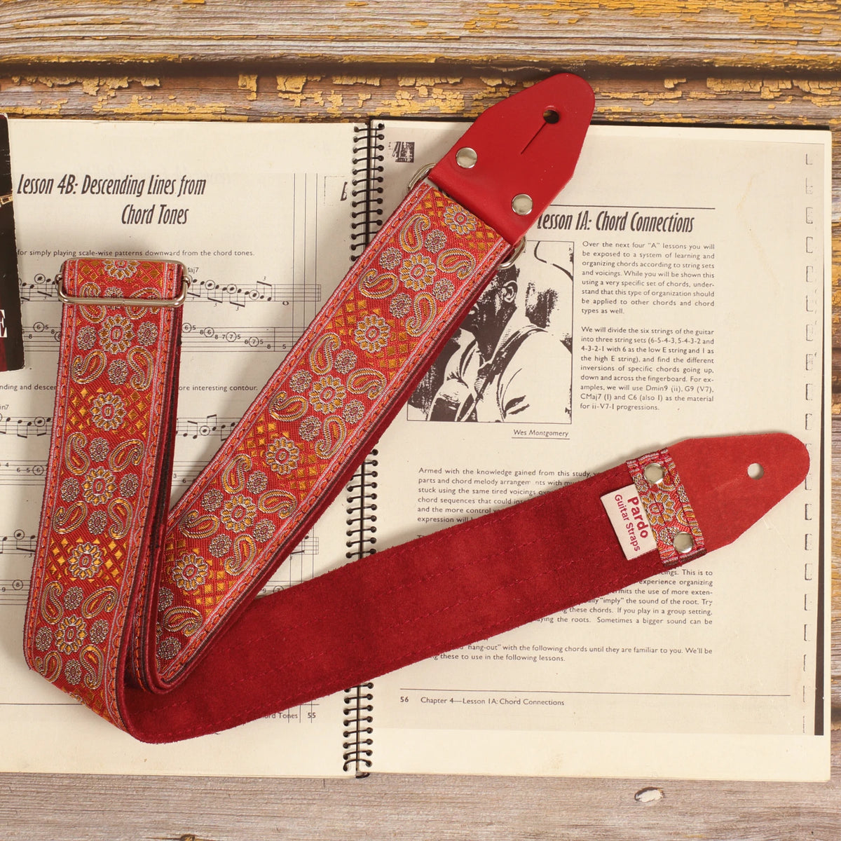 Red Andromeda guitar strap laid over a sheet music book, showing its red and orange jacquard pattern and burgundy suede backing.