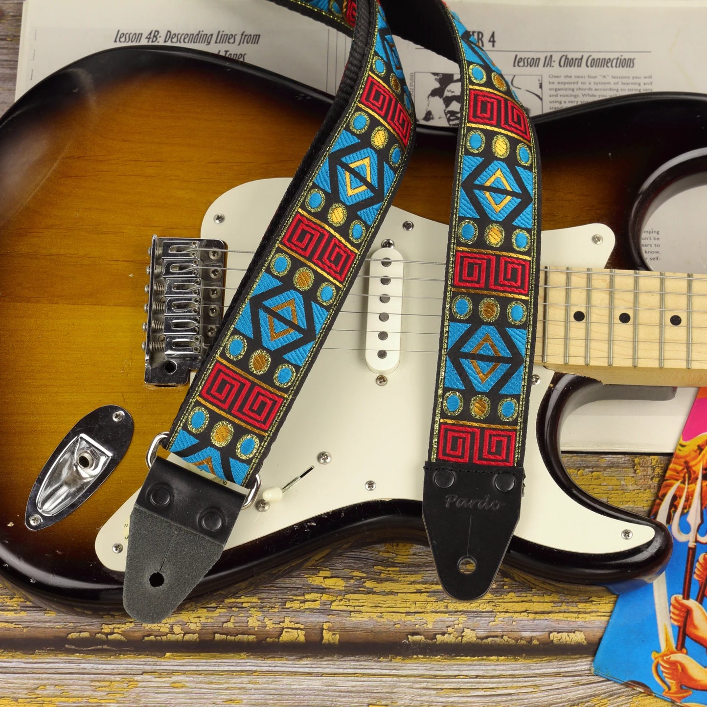 Sunstone Circuit guitar strap displayed on a sunburst Fender Stratocaster, showing its turquoise, red and golden geometric pattern.