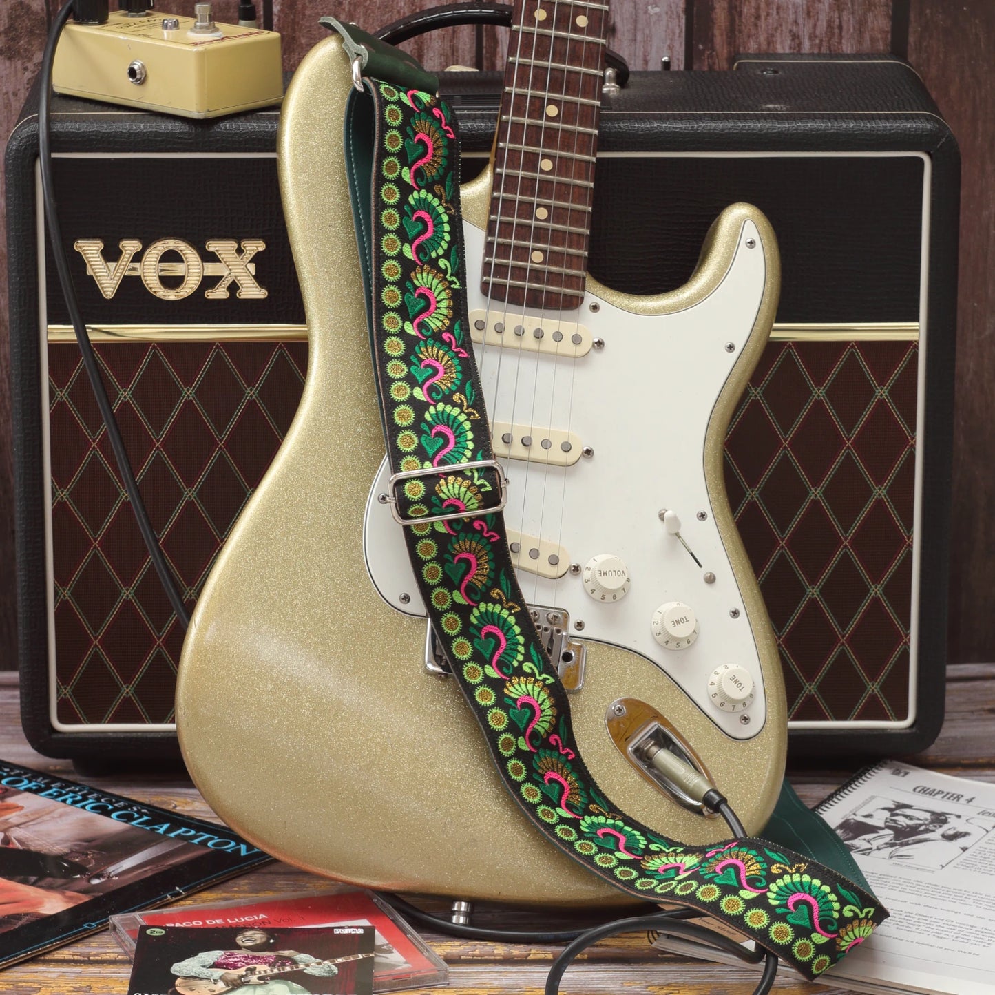 Swann Green guitar strap displayed beside a Fender Stratocaster, showing its green, pink and golden woven design on a black background.