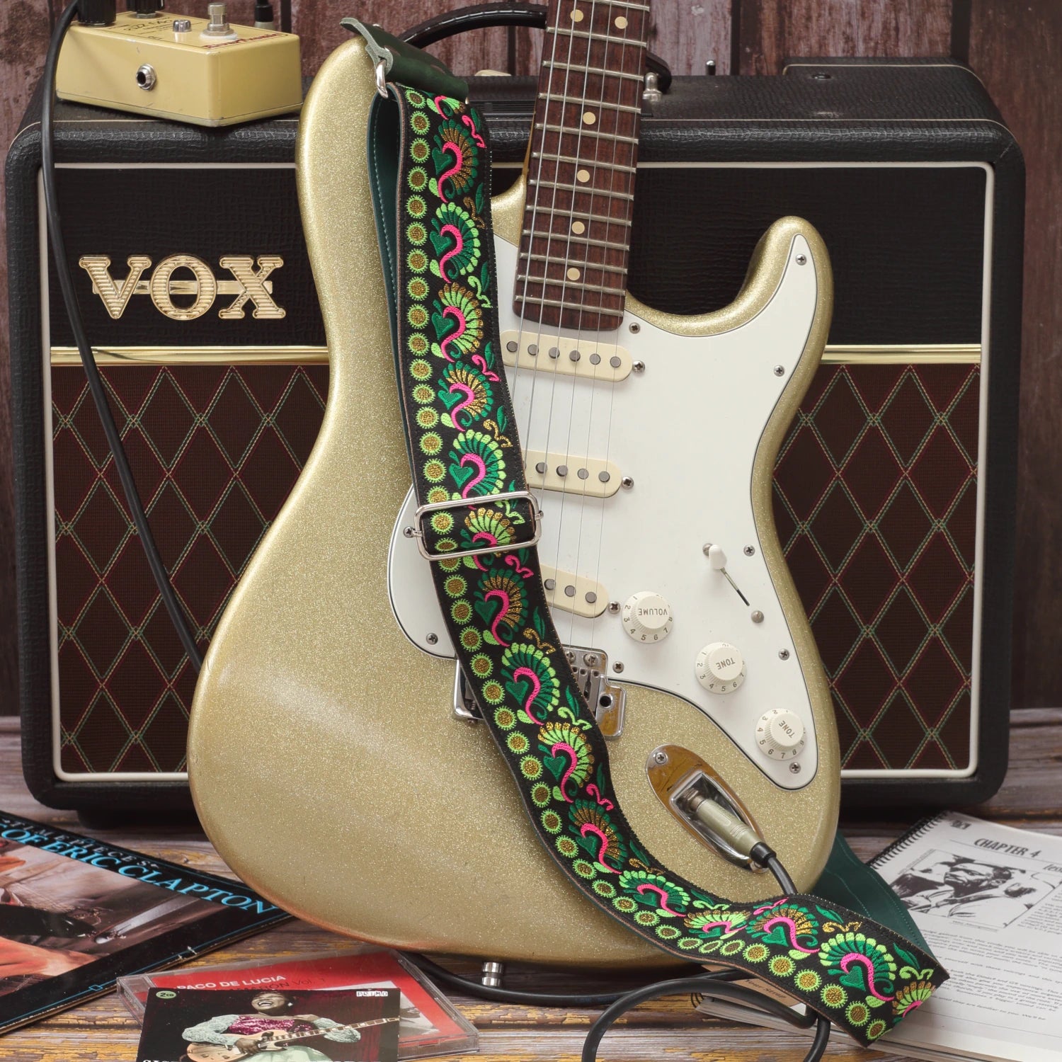Swann Green guitar strap displayed beside a Fender Stratocaster, showing its green, pink and golden woven design on a black background.