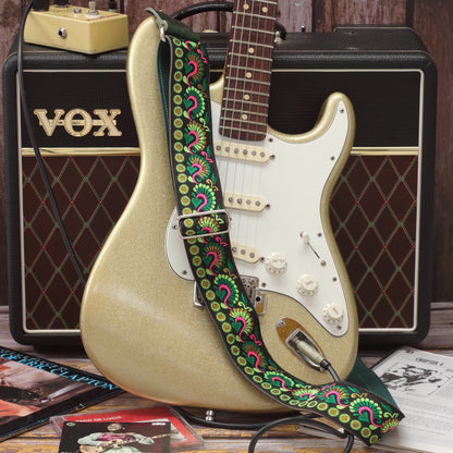 Swann Green guitar strap displayed beside a Fender Stratocaster, showing its green, pink and golden woven design on a black background.